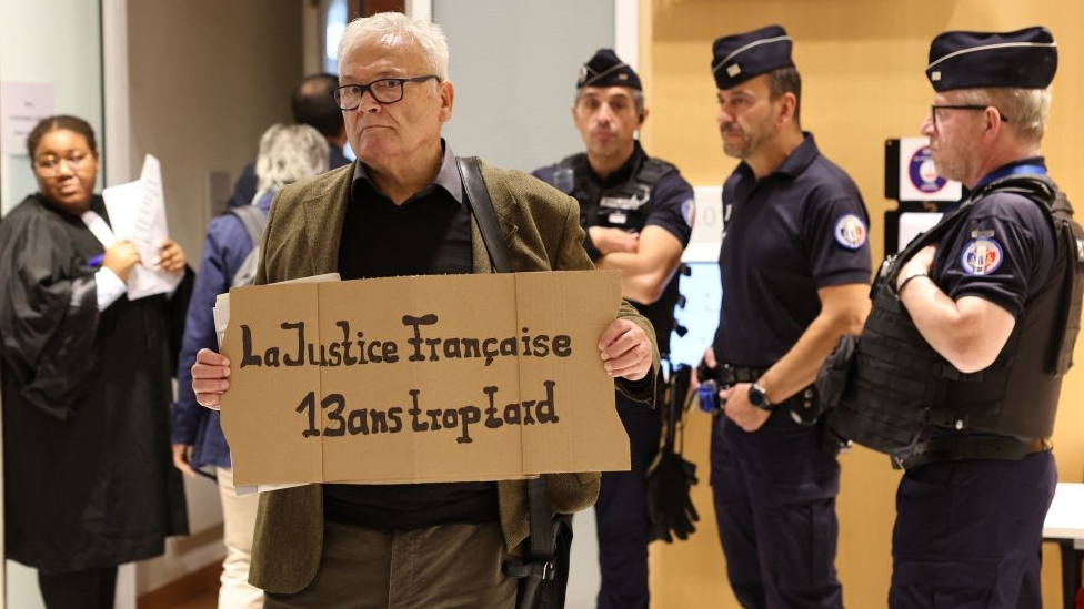 Один из посетителей суда держит плакат с надписью "Французское правосудие, опоздавшее на 13 лет", 10 октября 2022