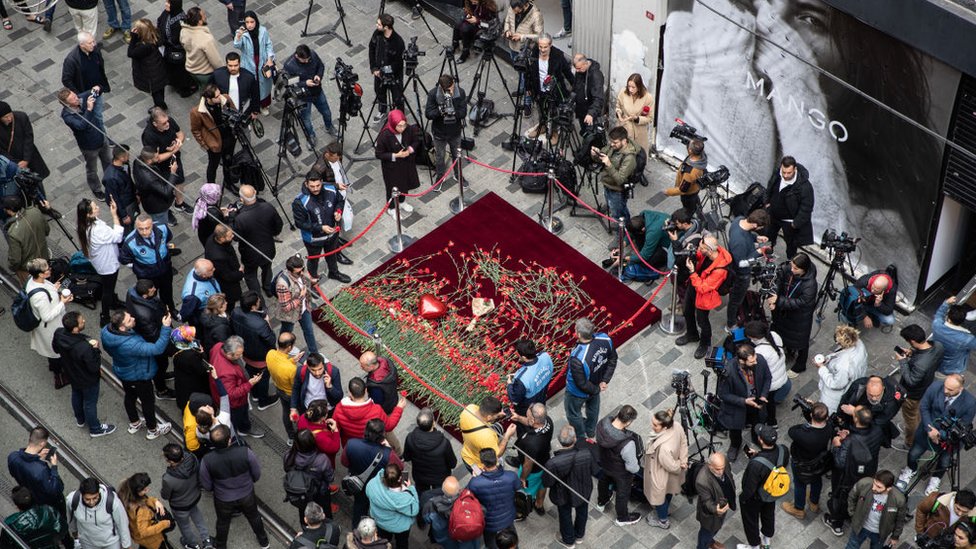 Красный ковер на месте взрыва, на котором лежат красные цветы, вокруг стоят люди