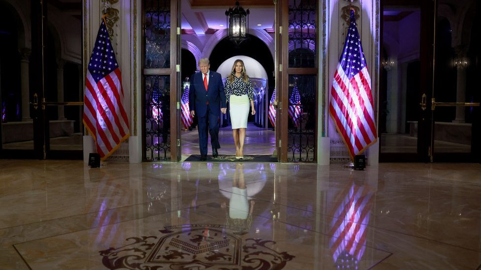 Дональд и Мелания Трампвыходят из стеклянных дверей, обрамленных американскими флагами.
