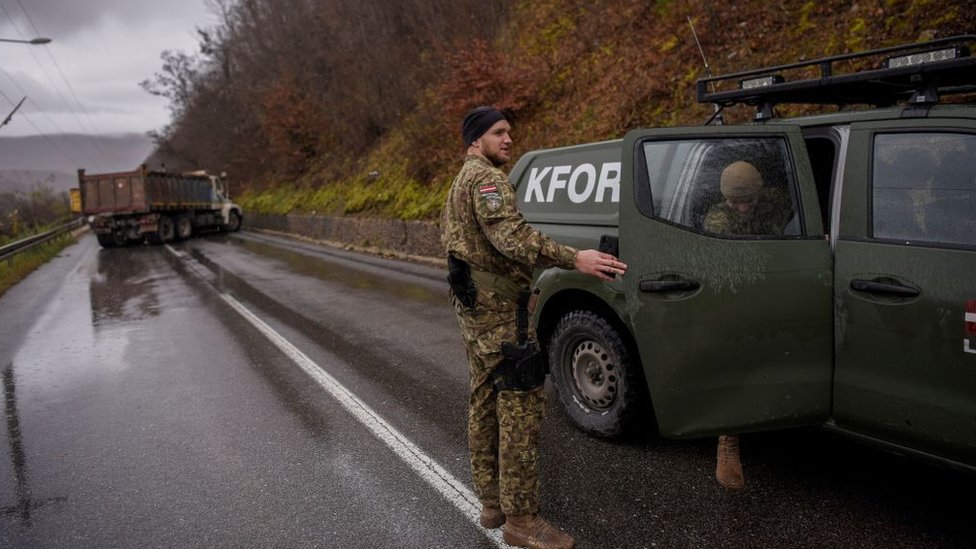 Солдаты около машины с буквами KFOR на дороге, перекрытой грузовиками