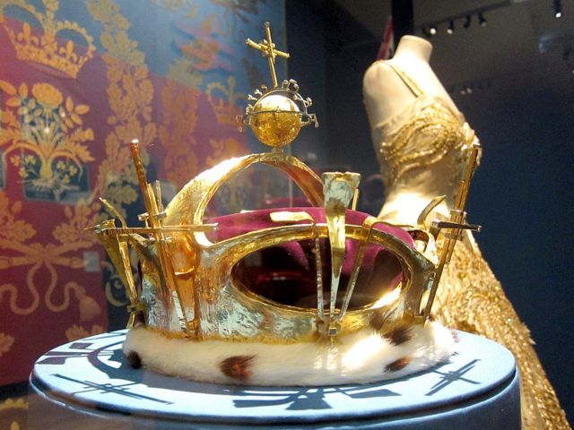Золотая корона под стеклом рядом с женским вечерним платьем